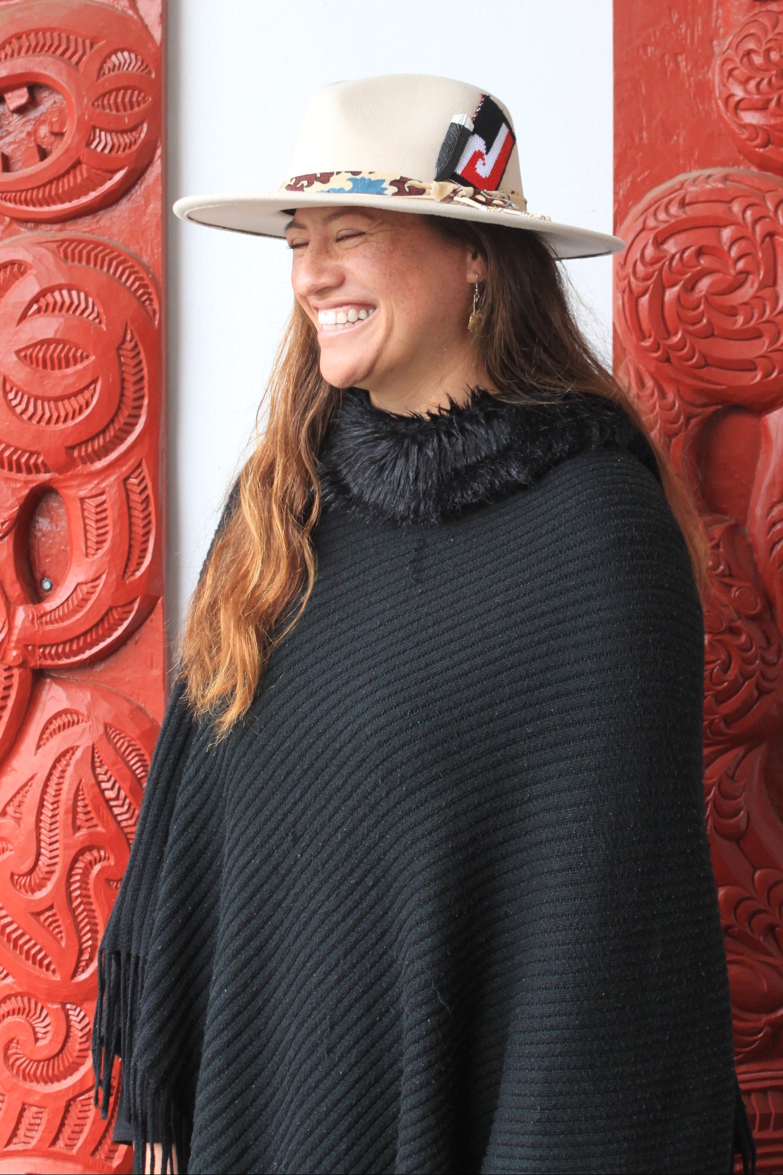 Person wearing a black coat and white hat leaning against a red decorative wall with intricate patterns.