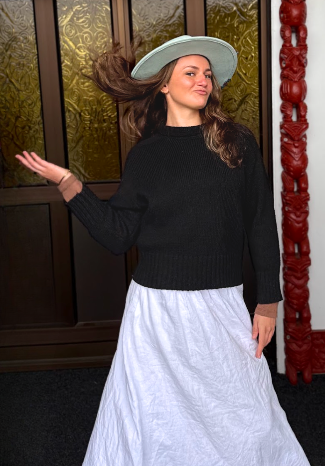 Woman wearing a black sweater and white skirt with a hat, standing in front of a decorative wall.